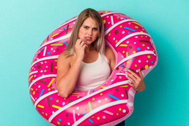 Şişme donutlu genç Rus kadın mavi arka planda izole edilmiş tırnak yiyor, gergin ve çok endişeli..