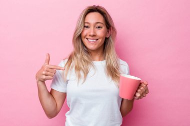 Genç Avustralyalı bir kadın pembe arka planda izole edilmiş pembe bir kupa tutuyor tişört fotokopi alanını işaret ediyor, gururlu ve kendinden emin.