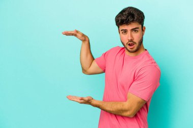 Mavi arka planda izole genç beyaz adam şok ve hayret içinde elleri arasında bir kopya boşluk tutuyor.