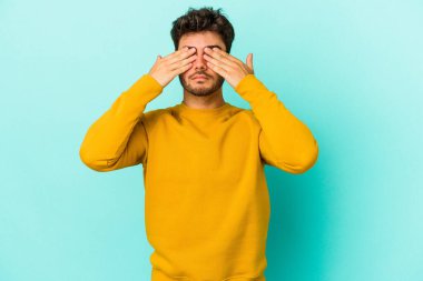 Mavi arka planda izole edilmiş genç beyaz adam elleriyle gözlerini kapatmaktan korkuyor..
