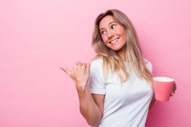 Pembe kupa tutan Avustralyalı genç bir kadın pembe arka planda izole edilmiş, başparmak uzakta, gülen ve kaygısız..