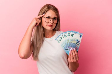 Elinde pembe arka planda izole edilmiş banknotlar tutan genç Rus kadın işaret parmağıyla yaptığı hayal kırıklığı jestini gösteriyor..