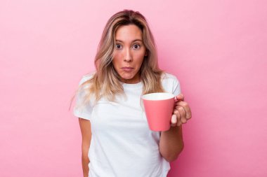 Pembe arka planda izole edilmiş pembe bir kupa tutan genç Avustralyalı kadın omuzlarını silker ve gözleri açık kalır..