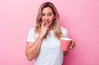 Pembe arka planda izole edilmiş pembe kupa tutan genç Avustralyalı kadın tırnaklarını yiyor, gergin ve çok endişeli..