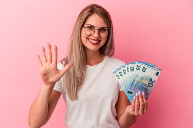 Pembe arka planda izole edilmiş banknotları tutan genç Rus kadın gülümseyerek beş numarayı parmaklarıyla gösteriyor..