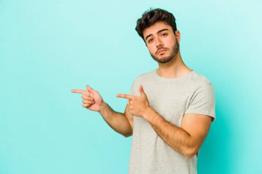 Mavi arka planda izole edilmiş genç beyaz adam işaret parmağıyla fotokopi alanını işaret ederek şok oldu..