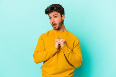Kafkasyalı genç adam mavi arka planda izole edilmiş şans için dua ediyor, şaşırmış ve ağzını açıp önüne bakıyor..