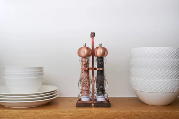 Old fashioned copper salt and pepper grinder set with Himalayan sea salt and peppercorns on a wooden shelf next to dishes and bowls