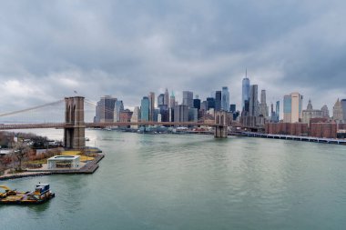 Brooklyn Köprüsü 'nün arka planda bulutlu bir gökyüzünün altında yüksek binalardan oluşan bir silueti olan bir nehir üzerinde uzanan görüntüsü.