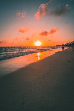 Güneş batarken, kıyı şeridi boyunca el ele yürüyen bir çift, suya sıcak bir parıltı saçıyor. Ayak izleri kumda görünür, palmiye ağaçları da kumsalda..