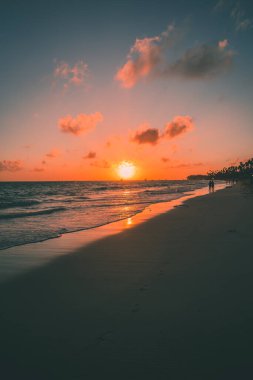 Güneş batarken yumuşak dalgalar kıyıya vurur ve gökyüzünde sıcak renkler bırakır. İnsanların siluetleri kumlu sahilde geziniyor, huzurlu anın tadını çıkarıyorlar..