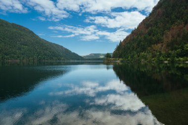 A serene lake showcases reflections of clouds and trees on its glassy surface, surrounded by majestic mountains under a bright blue sky on a fine day.
