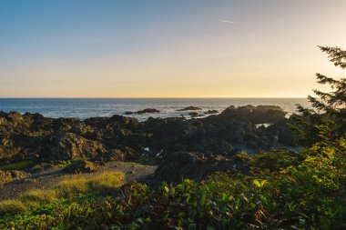 A beautiful coastline scene features rocky formations and gentle waves during sunset. The sky is warm with hues of orange and gold reflecting on the water.