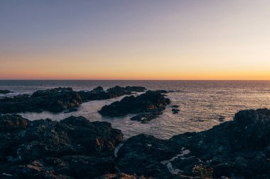 A beautiful sunset casts warm colors over the rocky coastline, with the ocean gently lapping against the shore, creating a serene atmosphere.