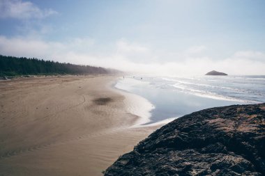 Soft waves lap at the sandy shore as surfers ride the water. A rocky outcrop contrasts with the smooth beach and trees in the distance on a sunny day.