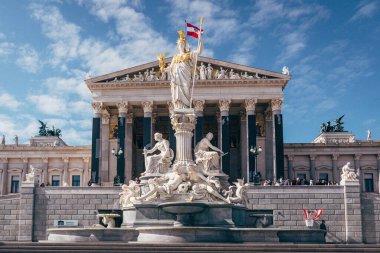 Ziyaretçiler, gündüz saatlerinde açık mavi bir gökyüzünün altında tarihi bir parlamento binasının önünde bayraklı büyük bir heykele hayran kalıyorlar..