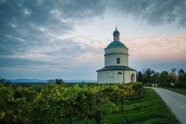 Alacakaranlıkta renkli bir gökyüzünün altında yemyeşil üzüm bağlarının arasında yuvarlak bir bina bulunur. Dönen bir patika tarlaların arasından geçiyor. Arka planda kayan tepeleri ve ağaçları gösteriyor..