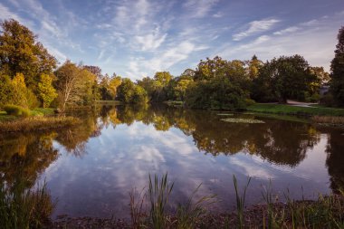 A tranquil lake reflects the blue sky and trees during sunset, showcasing autumn colors along the shore and inviting serenity.