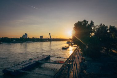 Güneş nehrin üzerinde batıyor, suya sıcak bir ışık tutuyor. İnsanlar kıyıda geziniyor, akşam atmosferinin tadını çıkarıyorlar..