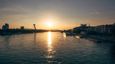 Güzel günbatımı nehir üzerinde sıcak renkler bırakır, şehir binaları ve kıyıda görünen bir köprü. İnsanlar huzurlu sahnenin tadını çıkarıyor..