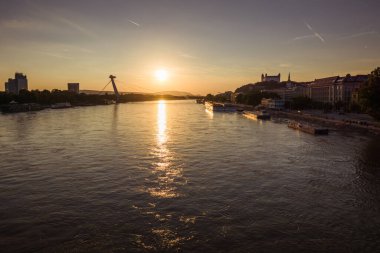 The sun sets over the Danube River, casting a golden glow on the water with boats lining the shore in Bratislava.