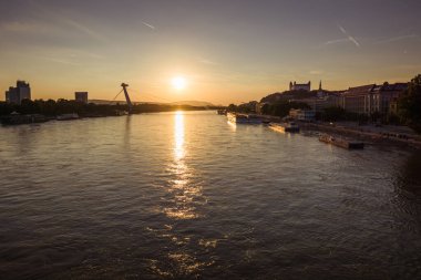 A beautiful sunset casts warm colors over the Danube River as boats float quietly. The cityscape of Bratislava is visible in the background.