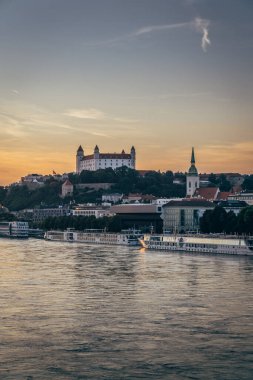 Bratislava Şatosu, güneş batarken görkemli bir şekilde bir tepenin üzerinde duruyor. Tuna Nehri 'nin ve şehir binalarının üzerine sıcak tonlar döküyor..