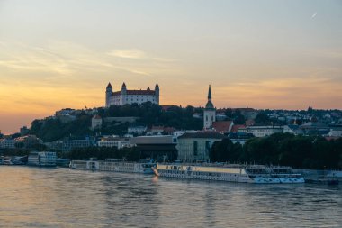 Günbatımı, Bratislava 'nın tepedeki şatosunu ve aşağıdaki nehirde yüzen teknelerini aydınlatıyor. Huzurlu bir manzara yaratıyor..