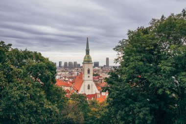 Tall church tower rises above red rooftops in a city skyline, surrounded by lush green trees under a cloudy sky.