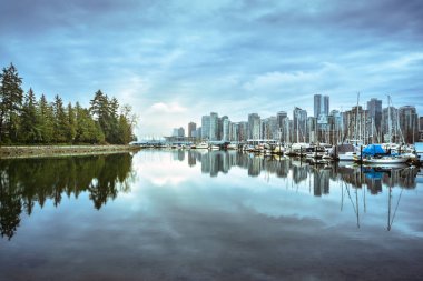 Uzun binalar bulutlu bir gökyüzüne karşı duruyor, teknelerin demirlediği durgun suya yansıyor. Ağaçlar kıyıya sınır çiziyor, alacakaranlıkta huzurlu bir manzara yaratıyor..