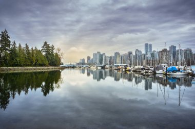 Yüksek binalar, sakin suya yansıyan çarpıcı bir ufuk çizgisi oluşturur. Uzun ağaçlarla çevrili tekneler kıyıya demir atar ve sakin bir akşam manzarası yaratır..