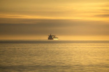 Bir balıkçı teknesi sersemletici bir günbatımında sakin sularda yüzeye sıcak renkler yansıtarak süzülür. Sahne, denizin dinginliğini yakalar..