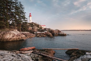 A lighthouse towers over rugged rocks by the sea at sunset. Calm waters reflect the warm hues of the sky. Trees frame the scene, creating a tranquil coastal atmosphere.