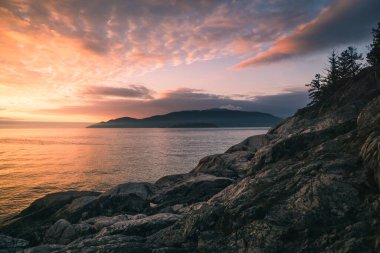 Bright orange and purple hues fill the sky as the sun sets over calm ocean waters. Rocky cliffs frame the landscape, with trees dotting the background on the distant shore.