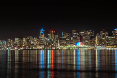 Şehirdeki uzun binalar gece gökyüzünü aydınlatır. Renkli ışıklar suyu yansıtır ve sakin bir atmosfer yaratır. Sahne, karanlık çöktükten sonra kentsel yaşamın güzelliğini yakalar..