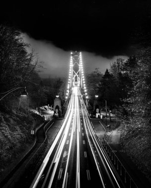 Parlak ışıklar gece köprüyü aydınlatır. Arabalar yolda uzun ışık yolları oluşturur. Etrafını saran ağaçlar ve karanlık bir gökyüzü dingin manzaraya derinlik katar..