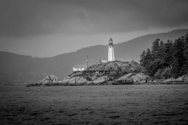 A lighthouse rises on a rocky island as dark clouds loom overhead. The surrounding sea reflects the white and gray tones of the sky, creating a moody atmosphere.
