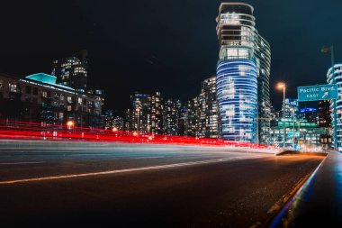 Parlak şehir binaları hareketli araçlardan gelen ışık çizgileriyle hareketli bir sokağı gece aydınlatıyor..