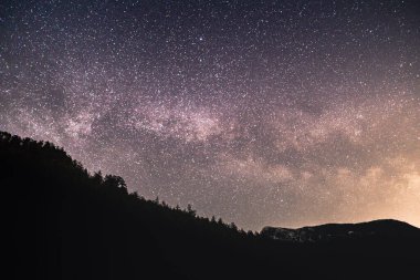 Stars fill the night sky above dark silhouettes of mountains, showcasing the beauty of the universe in a serene setting.