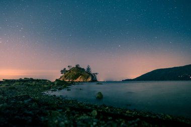 A tranquil coastal area features a small island under a starry sky, with calm waters reflecting distant lights from the shore.