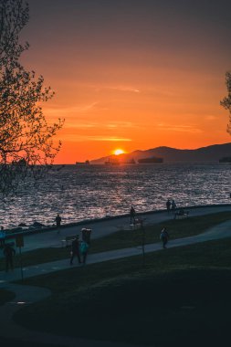 İnsanlar, güneş batarken kıyıya yakın yürürler. Sıcak renklerle gökyüzünü aydınlatırlar ve suya yansımalar bırakırlar..