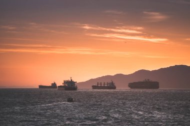 Günbatımında suda pek çok kargo gemisi görülebilir. Arka planda dağlar sakin bir atmosfer yaratır..