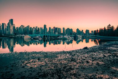 A vibrant sunset casts colors over a city skyline reflecting in the water. Boats are anchored along the shore, surrounded by urban landscape and a peaceful atmosphere.