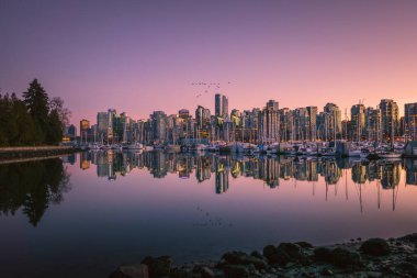 Uzun binalar güneş batarken Vancouver ufkunu aydınlatıyor ve suda renkli bir yansıma yaratıyor. Tekneler demirli, liman sahnesine cazibe katıyor..