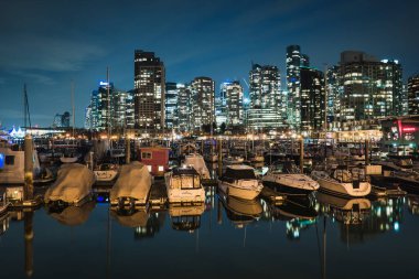Teknelerle dolu bir marinanın üstündeki parlak şehir binaları gece gökyüzünü aydınlatıyor. Su ufuk çizgisini yansıtıyor, alacakaranlıkta huzurlu ve canlı bir manzara yaratıyor..