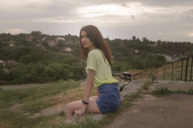 Kameraya bakan bir kızın düşünceli görüntüsü. Genç bir kız kaldırımda oturur ve yaz şehrine bakar. Manzara yüksek bir açıdan açılır. Kız geçenlerde bisiklete binmiş. Çok uzakta değil..