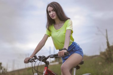 Genç bir kız bisikletini sürüyor. Kameraya bakıyorum. Bisiklet süren tişörtlü ve şortlu bir kız..