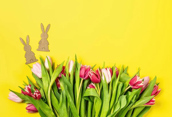 Pembe ve kırmızı laleli Paskalya kartı ve sarı arka planda ahşap tavşan.. 