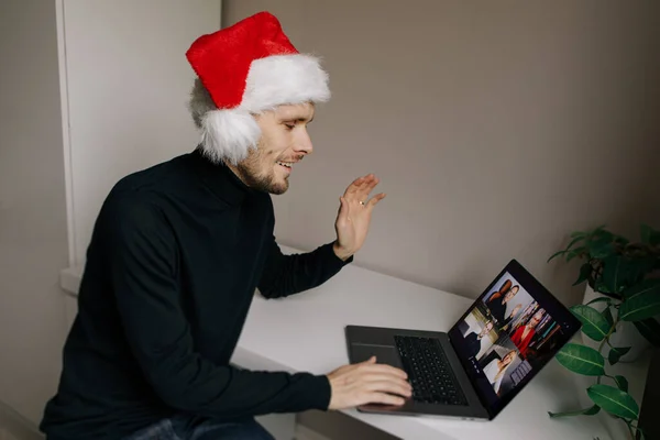 Remote Christmas video party. Family quality time during pandemic. Online video call conferencing. Young man wearing red Santa hat and holding a gift box. Virtual meeting conference calling from home