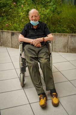 Yaşlı adam tekerlekli sandalyede oturuyor ve Covid-19 salgını sırasında maske takıyor.